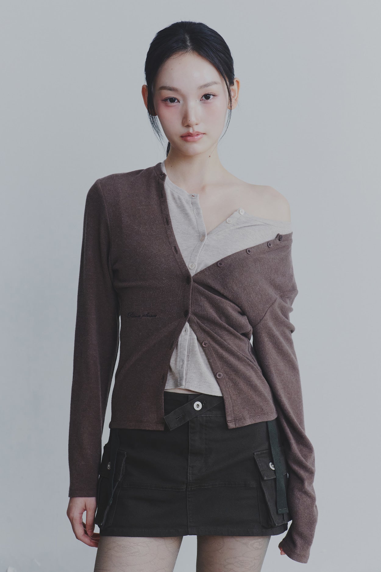 Woman wearing a brown cardigan over a beige top with a black skirt on a plain background