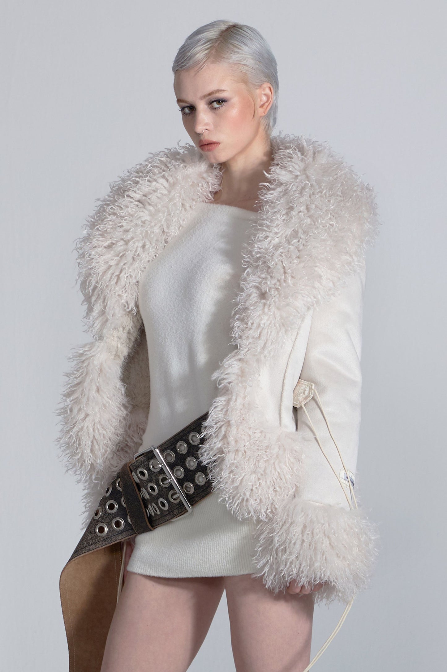 Woman wearing a white fur vest over a light-colored dress with a belt on a plain background