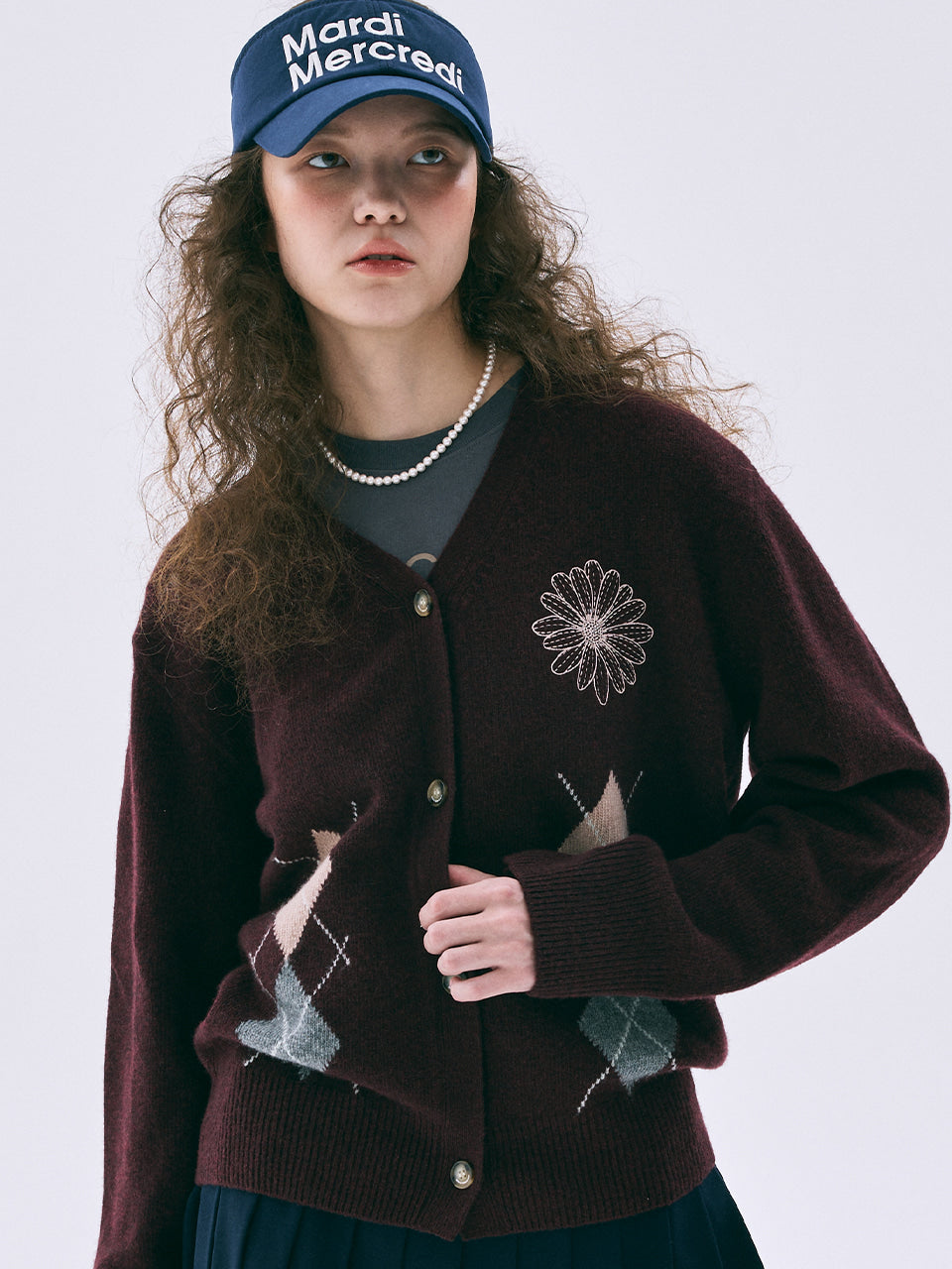 Woman wearing a brown cardigan with a floral design, dark blue cap, and pearl necklace on a white background