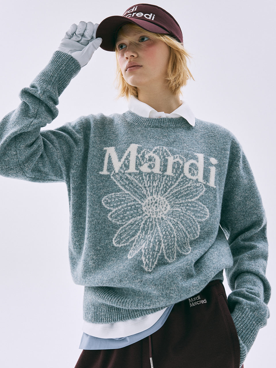 Person wearing a gray sweater with 'Mardi' and a flower design, holding a maroon cap.
