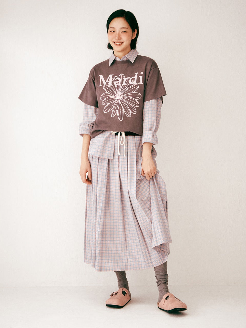 Person wearing a brown shirt with 'Mardi' print and plaid skirt on a white background