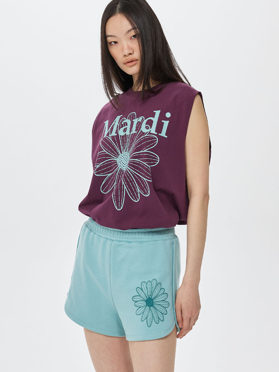 Woman wearing a purple sleeveless top with 'Mandi' and a flower design, paired with light blue shorts with a similar flower design on a white background.