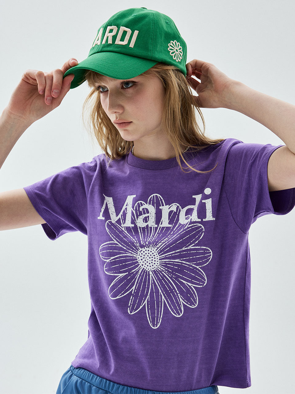 A young girl wearing a purple t-shirt with a white 'Mardi' and flower graphic, a green baseball cap with 'ARDI' text, and blue shorts, posing against a light gray background.