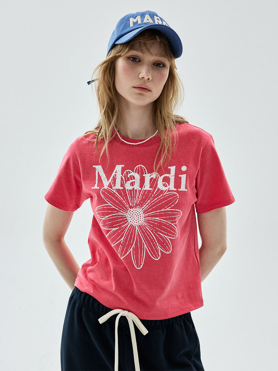 A model wearing a coral pink t-shirt with a white 'Mardi' and flower graphic, paired with a blue baseball cap and dark pants, against a light gray background.