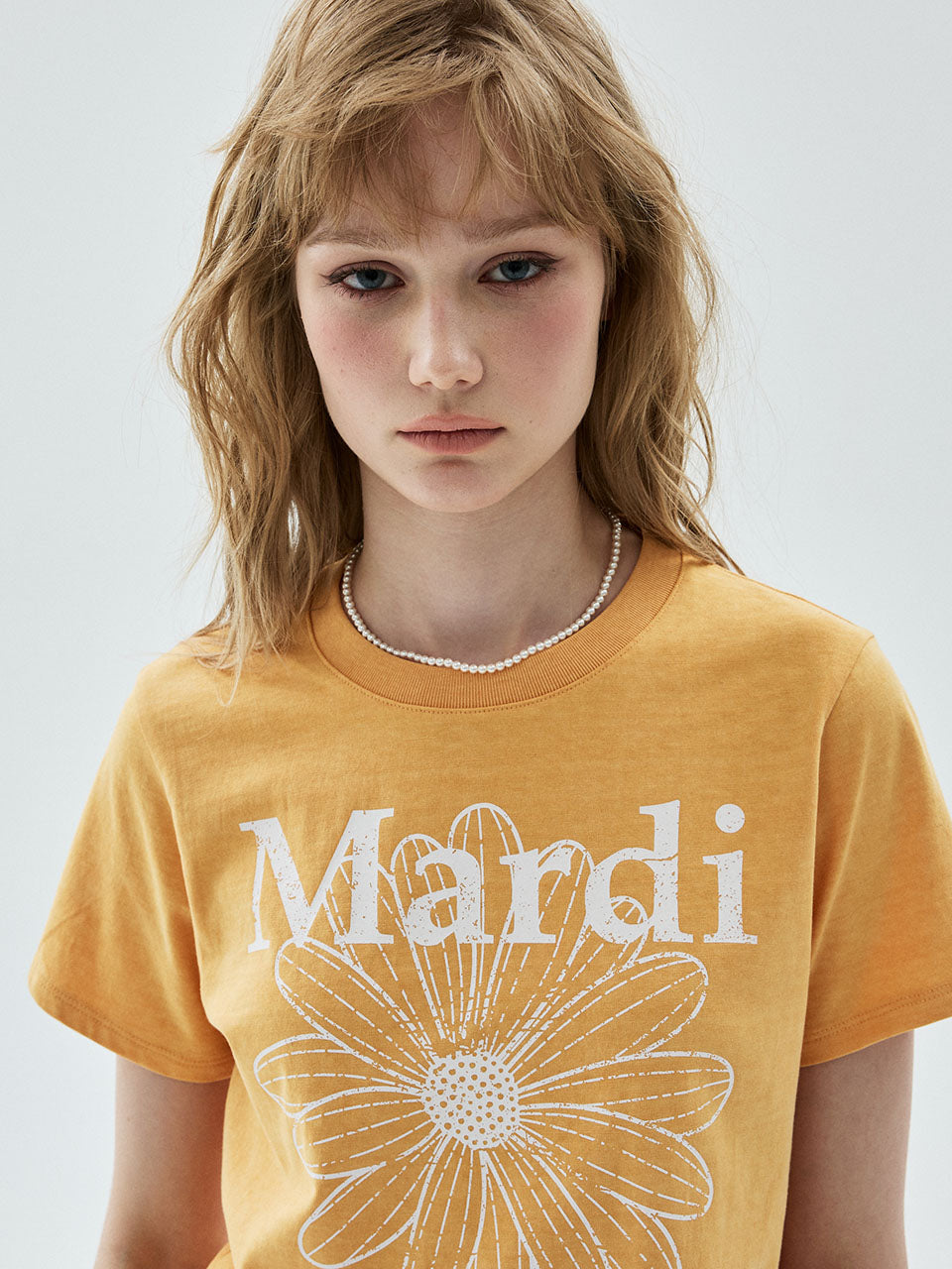 A woman wearing a mustard yellow graphic t-shirt with 'Mardi' and a daisy design printed in white on the front, set against a plain light gray background.