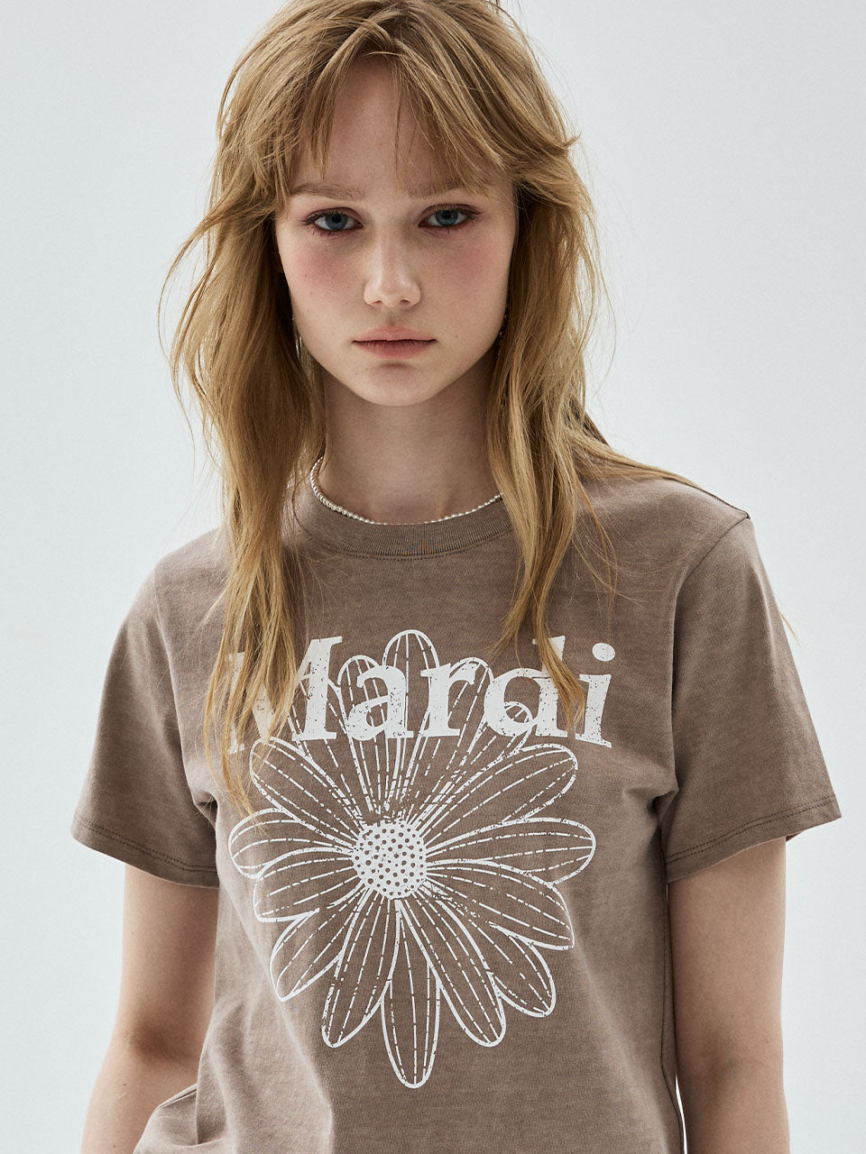 A woman wearing a brown short-sleeve t-shirt with a white graphic print of a flower and the word 'Mardi' across the chest, set against a plain light gray background.