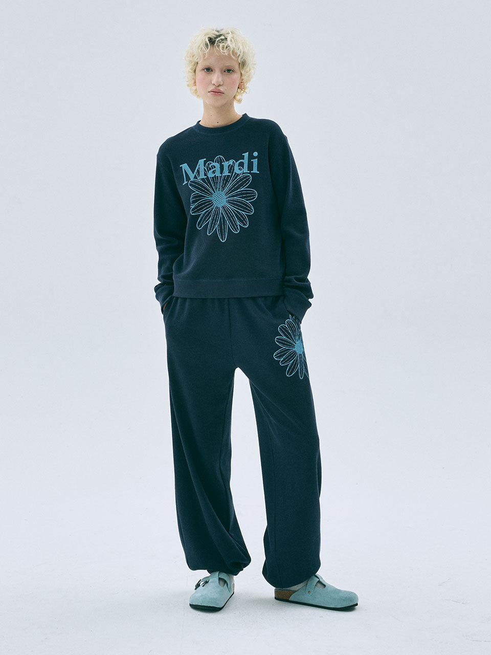 Person wearing a dark green sweatshirt and pants with 'Mardi' and floral designs on a light gray background