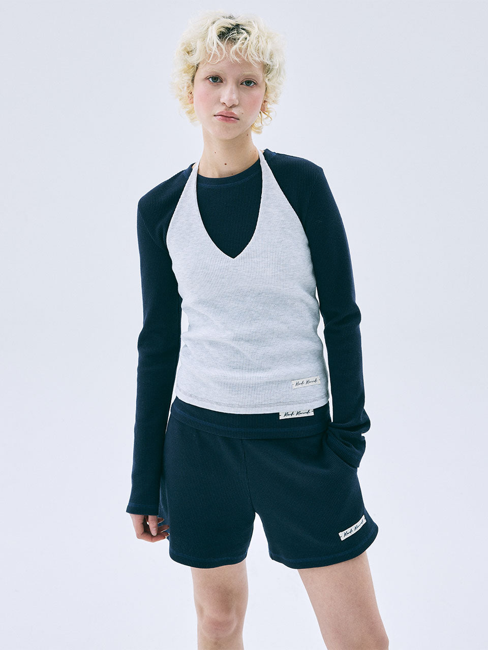 Person wearing a navy long-sleeve shirt over a white tank top and dark shorts on a light gray background