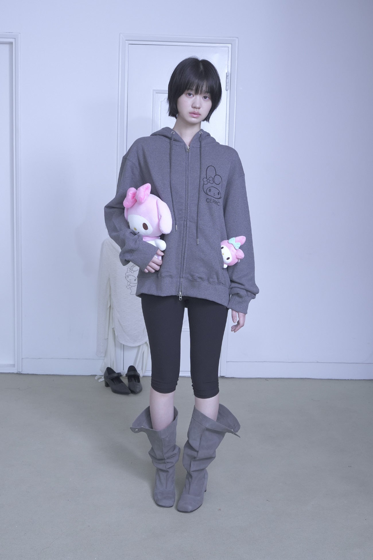 Person wearing a gray hoodie with cartoon character design, holding plush toys, standing against a plain background.