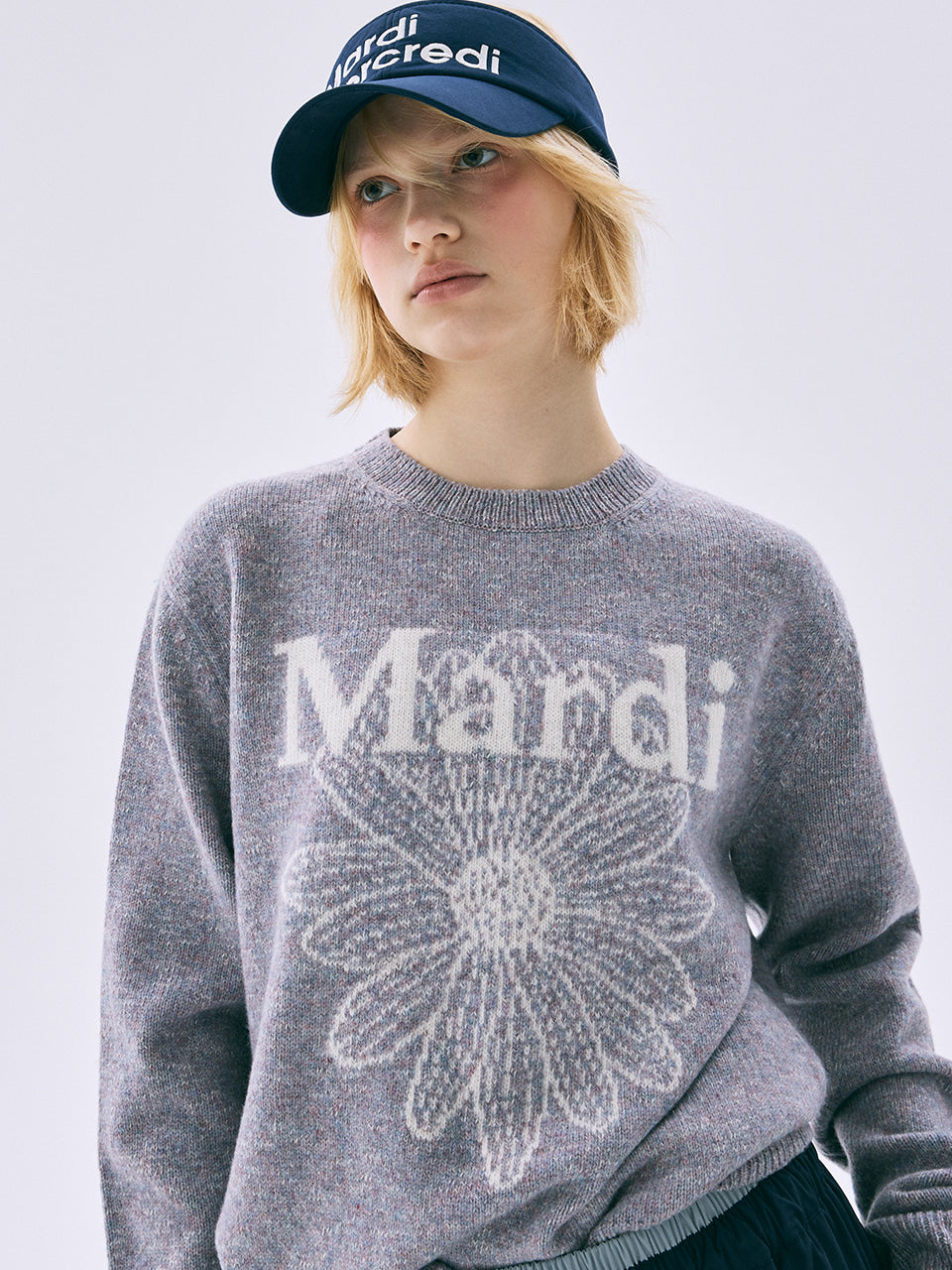 Person wearing a gray sweater with 'Mardi' text and a blue cap on a light background